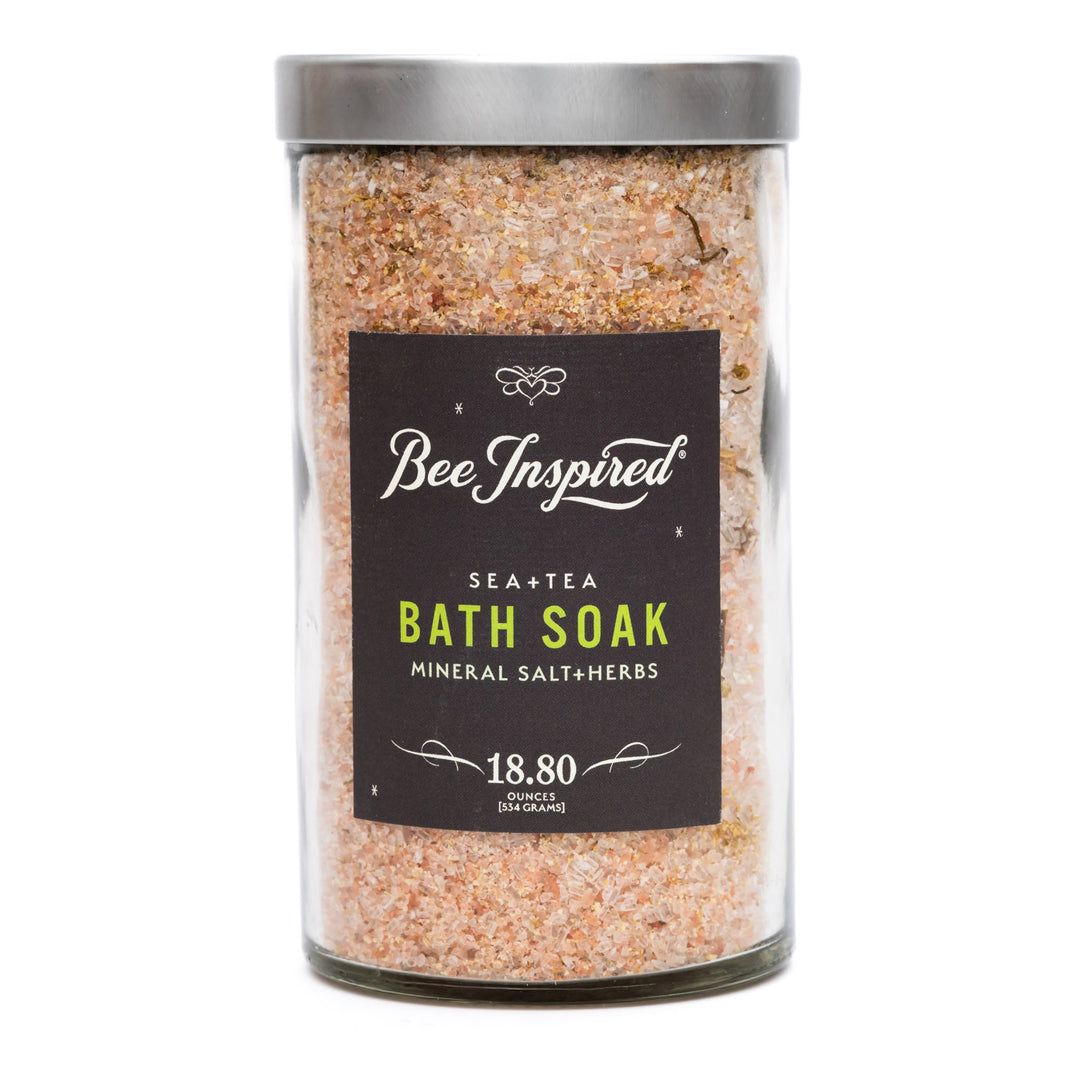 A clear glass jar filled with pinkish-beige mineral salt bath soak, topped with a silver lid and featuring a black label that reads 'Bee Inspired' and 'Sea+Tea Bath Soak'.