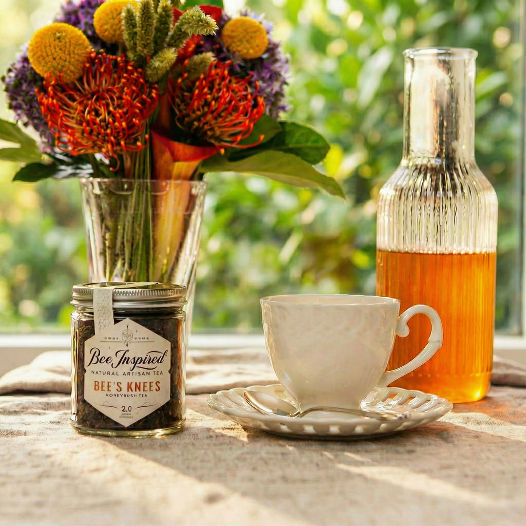 Tea cup with saucer, jar of 'Bee's Knees' tea, and bottle of honey on a wooden table with flowers in the background.