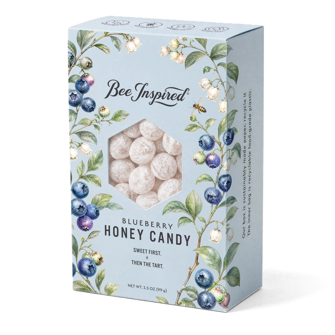 A light blue rectangular box of Bee Inspired Blueberry Honey Candy, featuring watercolor illustrations of blueberries and green leaves around a hexagonal window showing the round, off-white honey candies inside.