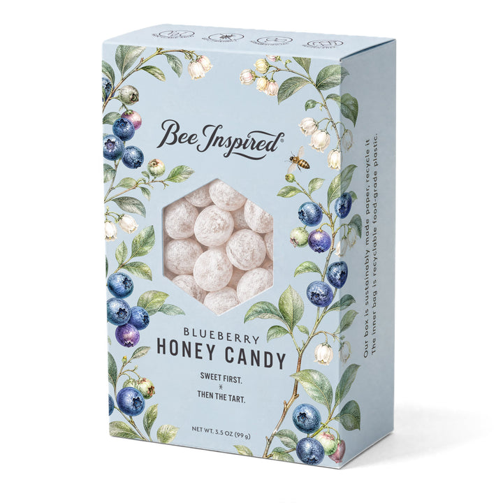 A light blue rectangular box of Bee Inspired Blueberry Honey Candy, featuring watercolor illustrations of blueberries and green leaves around a hexagonal window showing the round, off-white honey candies inside.