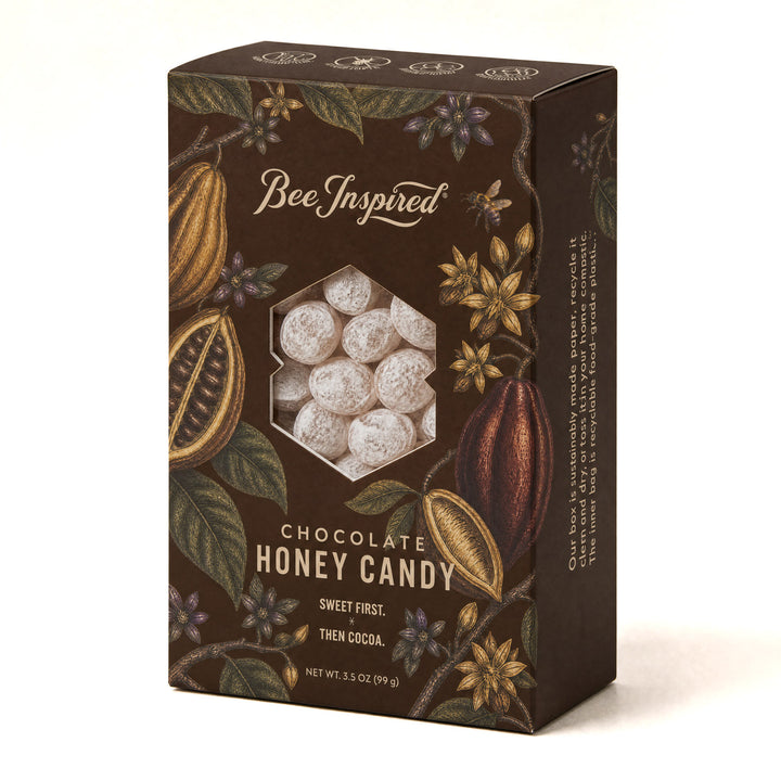 A box of Bee Inspired Chocolate Honey Candy, featuring a dark brown packaging design decorated with cocoa plants and a hexagonal window showing the white, frosted candy pieces inside.