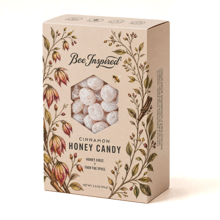A rectangular beige box of Bee Inspired Cinnamon Honey Candy, featuring a hexagonal window that reveals round, frosted white candies inside. The packaging is decorated with vintage-style botanical illustrations of flowers and leaves in green, brown, and red, with the brand name &