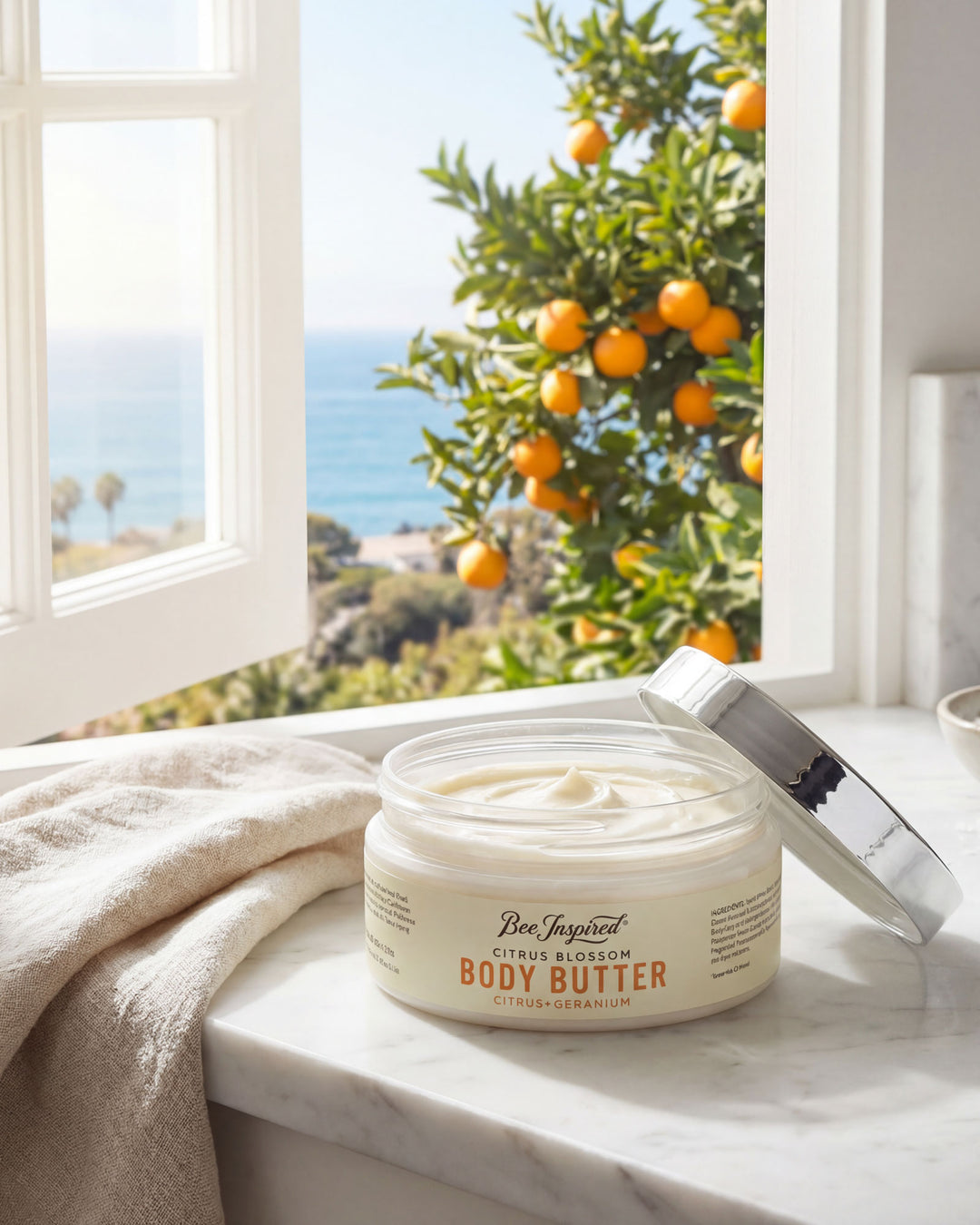 Jar of body butter on a marble surface with an orange tree and ocean view through a window.