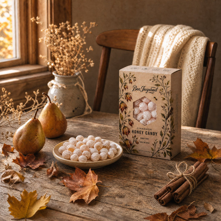 A styled tabletop scene showing a box of Ben Jussied Honey Candy next to a small bowl of sugar-dusted honey balls, with pears, autumn leaves, cinnamon sticks, and a cozy chair in the background.
