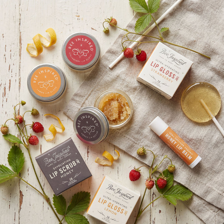 Collection of lip care products including lip gloss, lip balm, and lip scrub on a wooden surface with strawberries and leaves.