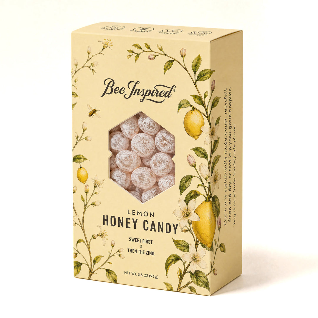 A box of Bee Inspired Lemon Honey Candy, featuring a pale yellow packaging decorated with illustrations of lemons, bees, and white flowers. A hexagonal window on the front reveals the round, frosted white candy inside.