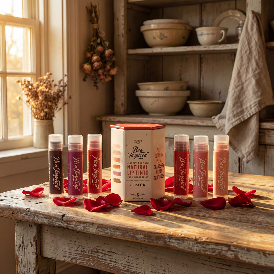 Set of lip balm tubes and a box on a wooden table with rose petals, in a rustic kitchen setting.