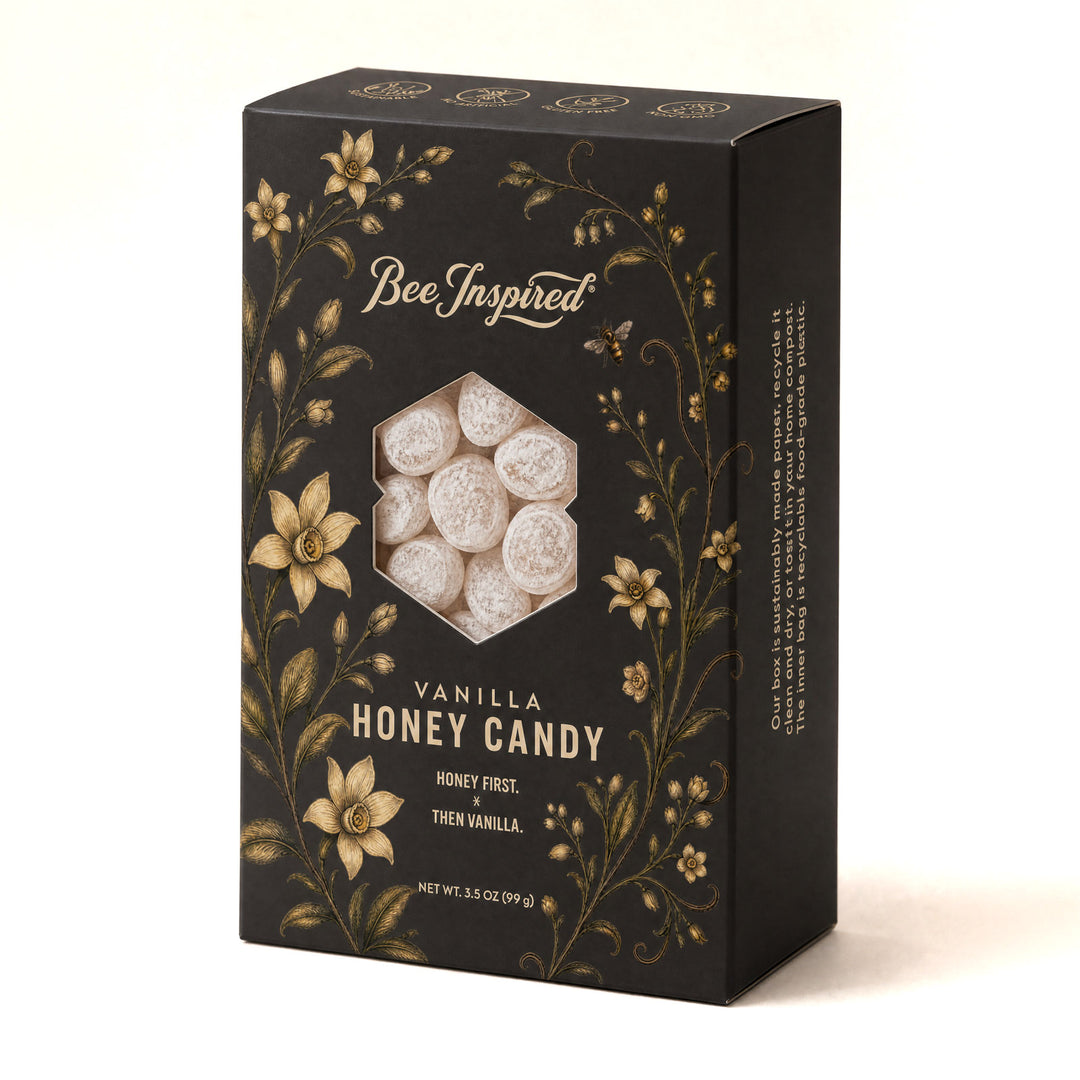 A rectangular black box of Bee Inspired Vanilla Honey Candy, featuring gold floral illustrations and a hexagonal window showing the white honey-coated candies inside, set against a neutral light beige background.